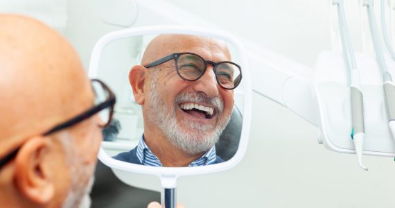 Happy senior patient admiring his perfect smile after a dental implant treatment, holding a mirror in his hand at the dental clinic
