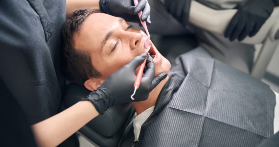 Man patient closed his eyes feeling fear while experienced dentist checking condition of his tooth and gums with special tools, providing high quality dental care in relaxing atmosphere