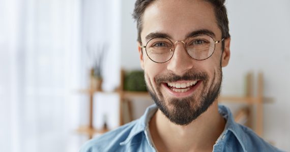 Close up shot of cheerful satisfied attractive male with stubble, has broad smile, wears round spectacles, rejoices success at work, stands against cozy interior. Fashionable designer glad be praised