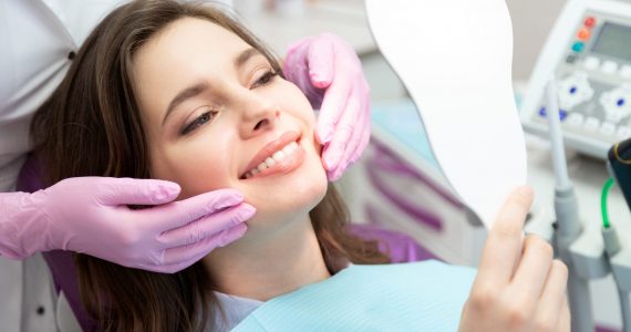 Happy patient after tooth treatment at dental clinic. Smiling girl with white well-groomed teeth after taking doctor help. Satisfied lady look at mirror enjoy healthy enamel after whitening procedure