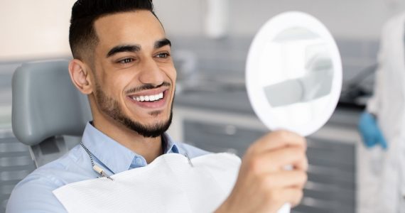 Happy Handsome Man Looking To Mirror After Teeth Treatment In Modern Clinic, Middle Eastern Male Patient Sitting In Chair In Stomatological Cabinet And Enjoying His New Smile, Closeup