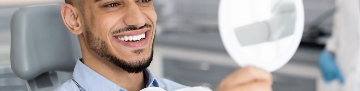 Happy Handsome Man Looking To Mirror After Teeth Treatment In Modern Clinic, Middle Eastern Male Patient Sitting In Chair In Stomatological Cabinet And Enjoying His New Smile, Closeup