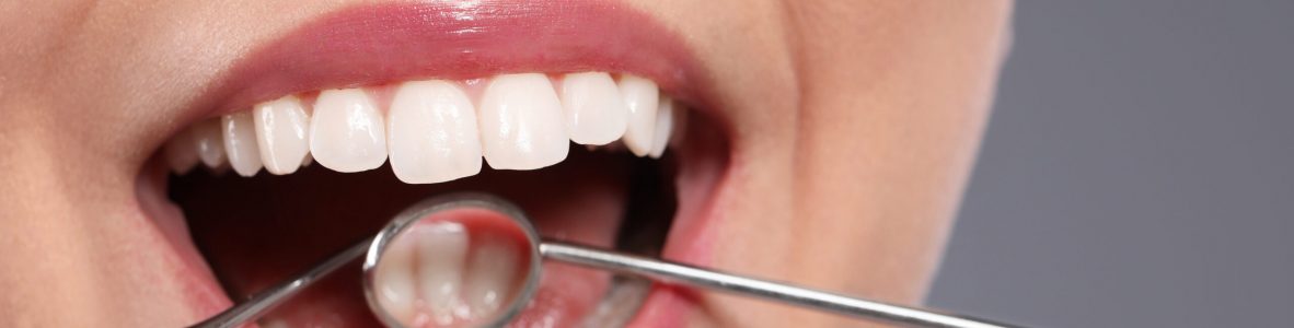 Examining woman's teeth with dentist's mirror on grey background, closeup