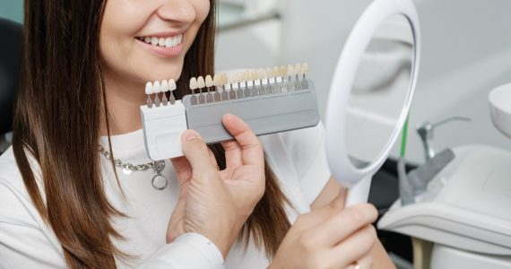 Young male dentist in white coat show veneers enamel color palette for beautiful patient woman sitting in a dental unit. Healthcare oral aesthetic treatment.