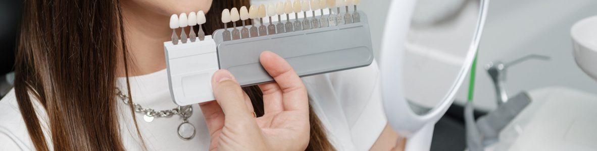 Young male dentist in white coat show veneers enamel color palette for beautiful patient woman sitting in a dental unit. Healthcare oral aesthetic treatment.