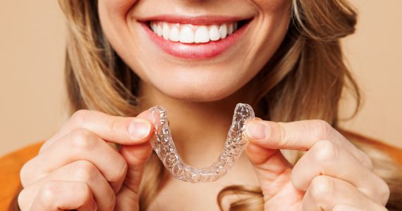 Close up cropped young woman wear orange shirt casual clothes hold in hand invisible transparent aligners, invisalign bracer smile isolated on plain pastel light beige background. Lifestyle concept