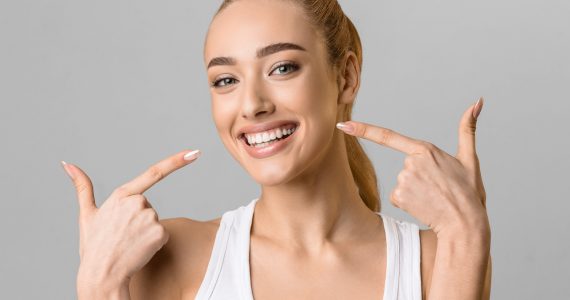 Orthodontic concept. Happy girl showing her beaming white teeth with two forefingers, grey background